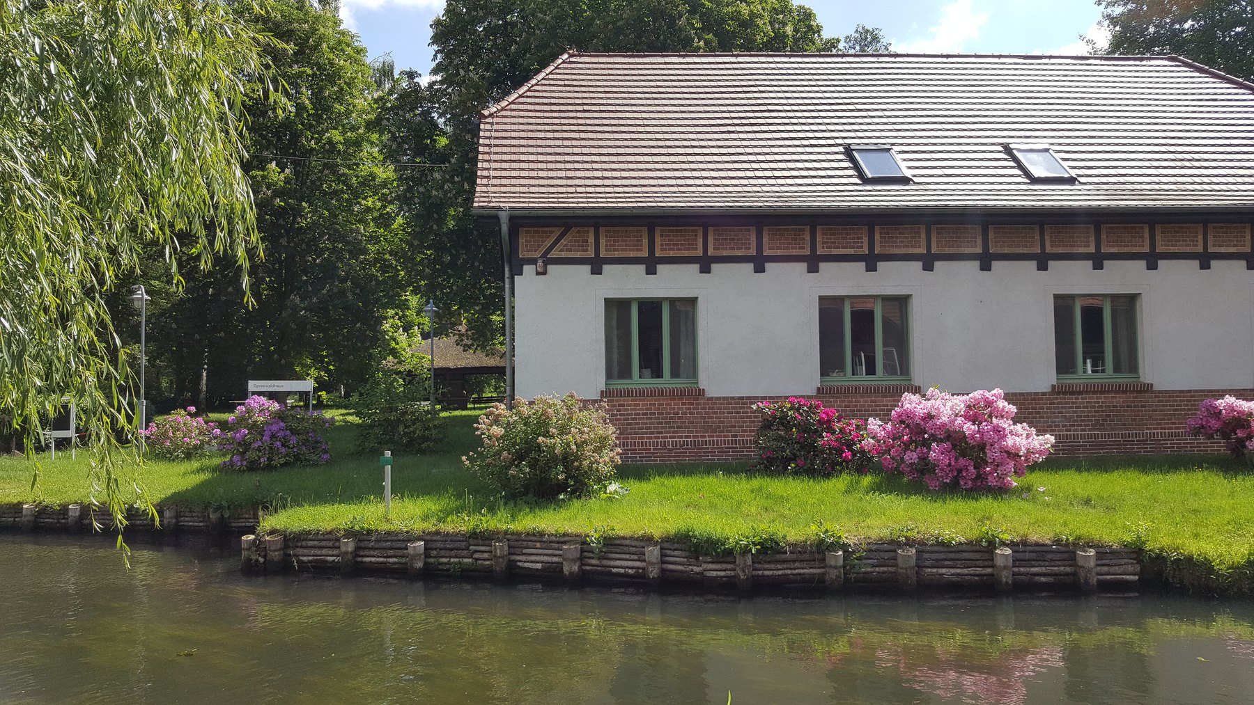 Das Apartmenthaus des Spreehofs vom Wasser aus gesehen, mit Wiese und blühenden Hortensien am Ufer